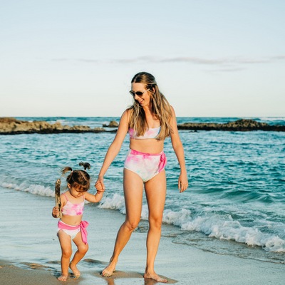 Mom and Daughter in Matching Cotton Candy Tie Dye Swimsuits.  The mom is in a high rise bikini bottom and a sporty bikini top.  The daughter has a matching bikini. 