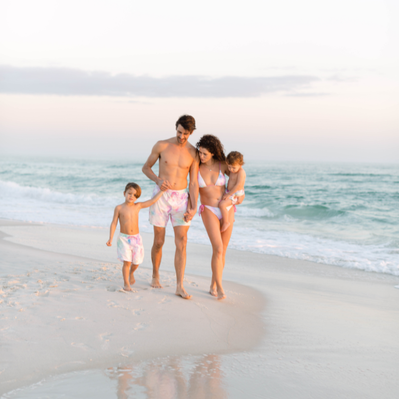 Mom, Dad and Two toddler sons walking on the beach in Florida wearing matching bathing suits.  Charlie Swims cotton candy tie dye collection is perfect for family Vacations. 
